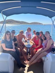 Group of friends smiling and relaxing on a covered pontoon boat around a small table on a sunny lake, sparkling blue water and low desert hills in the background.