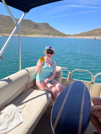 Person in a captain’s hat and sunglasses on a pontoon boat at a turquoise desert lake, wearing a light-blue swimsuit and holding a yellow spiral snack on a stick with cactus-studded hills and clear blue sky behind.