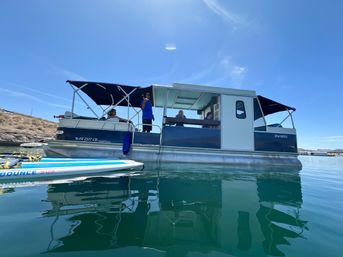 Pontoon boat with shaded canopies and people relaxing on a calm blue lake under a sunny sky, a paddleboard floating nearby with clear water reflections.