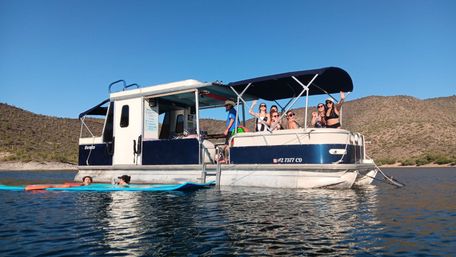 Group of people waving and lounging on a blue-and-white pontoon boat anchored on a calm desert lake, with paddleboards floating nearby and sunlit scrubby hills under a clear blue sky.