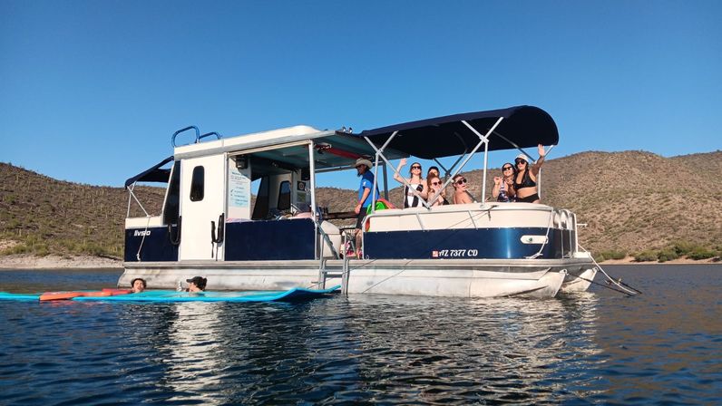 Group of people waving and lounging on a blue-and-white pontoon boat anchored on a calm desert lake, with paddleboards floating nearby and sunlit scrubby hills under a clear blue sky.