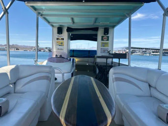 Pontoon boat interior with white cushioned benches, surfboard-style wood table, teal canopy and marina view across a calm lake