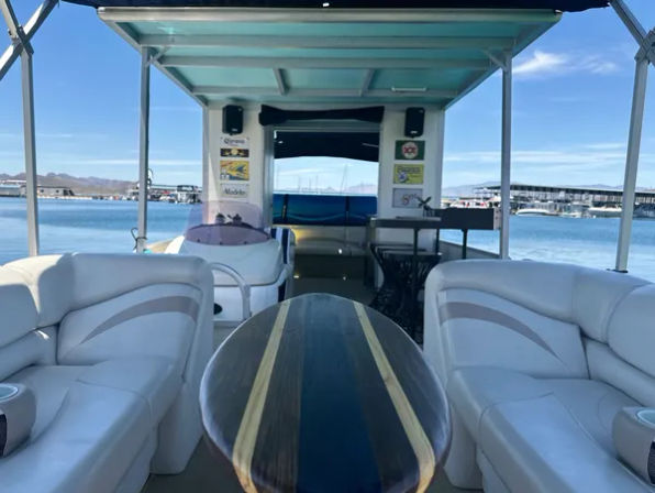 Pontoon boat interior with white cushioned benches, surfboard-style wood table, teal canopy and marina view across a calm lake