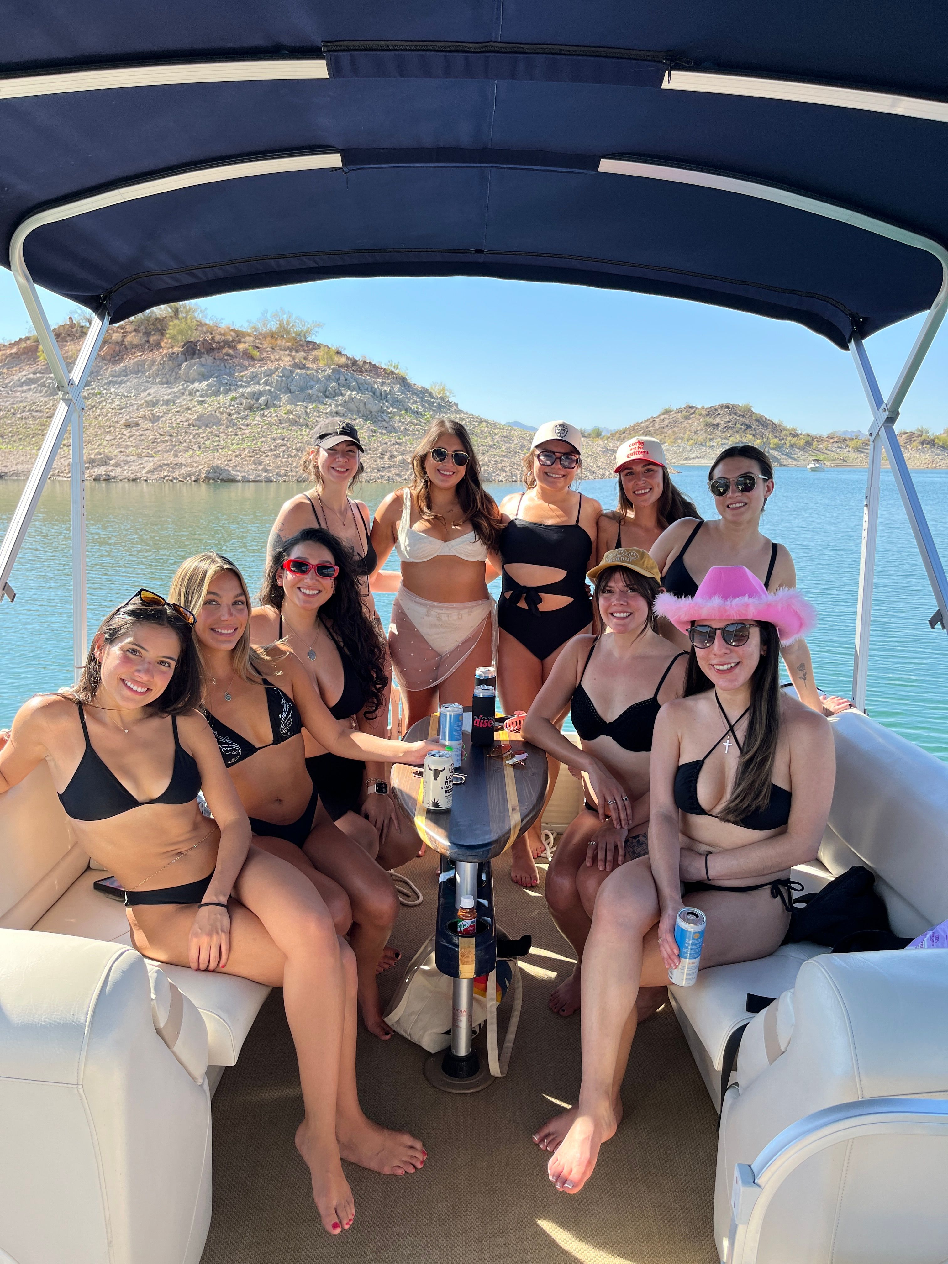 Group of women in bikinis lounging on a covered pontoon boat at a sunny desert lake with rocky shoreline, drinks on the table, sunglasses and hats.
