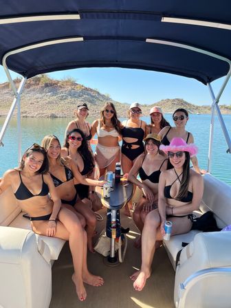 Group of women in bikinis lounging on a covered pontoon boat at a sunny desert lake with rocky shoreline, drinks on the table, sunglasses and hats.