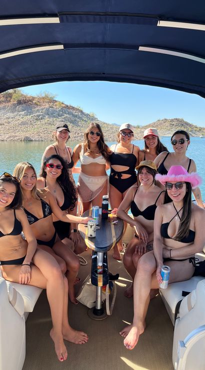 Group of women in bikinis lounging on a covered pontoon boat at a sunny desert lake with rocky shoreline, drinks on the table, sunglasses and hats.