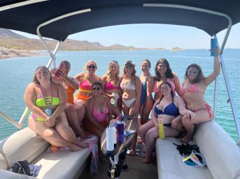 Group of friends in colorful swimsuits posing on a shaded pontoon boat over a clear blue lake with rocky desert hills in the background on a sunny day
