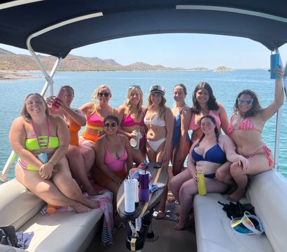 Group of friends in colorful swimsuits posing on a shaded pontoon boat over a clear blue lake with rocky desert hills in the background on a sunny day
