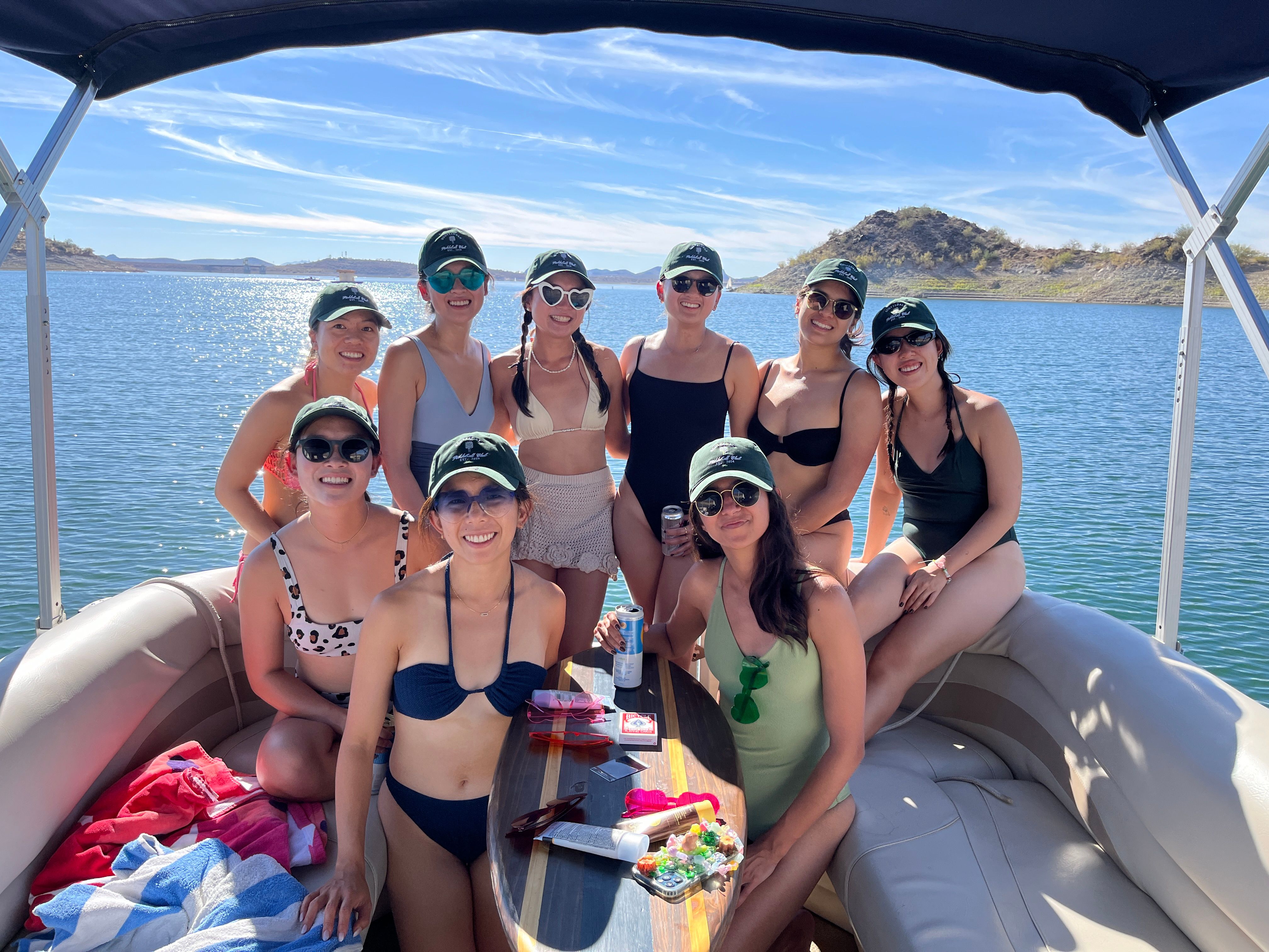 Eight women in swimwear and matching caps smiling on a pontoon boat during a sunny lake day with blue sky and low desert hills on the shoreline.