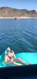Smiling man on a turquoise float in a blue desert lake, holding a drink and flashing a peace sign, with cactus-covered hills and a pink houseboat in the distance.