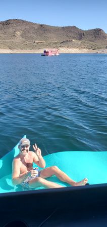 Smiling man on a turquoise float in a blue desert lake, holding a drink and flashing a peace sign, with cactus-covered hills and a pink houseboat in the distance.