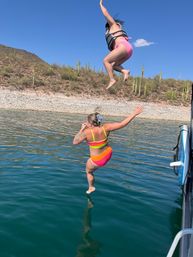 Two people jumping from a boat into a clear desert lake — one in a neon swimsuit and another in a pink swimsuit with a life jacket mid-air, rocky shoreline with tall cacti and bright blue sky.
