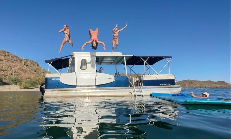 Three people in swimsuits mid-air diving off a pontoon boat into a clear lake, a paddler filming nearby, desert shoreline with cacti under a bright blue sky