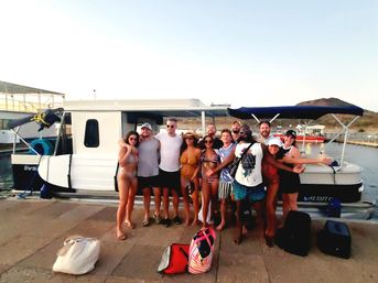 Smiling group of friends in swimsuits and shorts posing on a marina dock in front of a pontoon boat with bags on the pier, sunset beach-vacation vibe.
