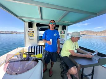 Two men relaxing on a covered pontoon boat at a sunny lake marina—one holds a canned drink while the other checks a phone at a small table, with docks and hills in the background.