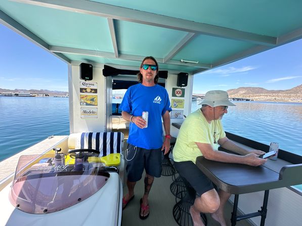 Two men relaxing on a covered pontoon boat at a sunny lake marina—one holds a canned drink while the other checks a phone at a small table, with docks and hills in the background.