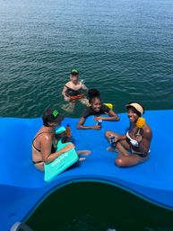 Four friends enjoying a sunny summer day on a bright blue floating dock in open water — two seated on the platform and two in the water — smiling and holding canned drinks and a tropical fruit skewer.