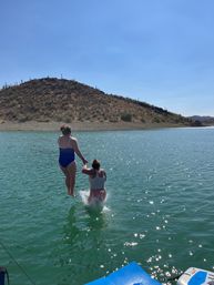 Two friends holding hands jumping from a dock into a sunlit desert lake with sparkling turquoise water, a cactus-dotted hillside and clear blue sky