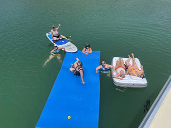 Friends floating on a bright blue water mat, a paddleboard and an inflatable lounger in a sunlit lake, sipping drinks and relaxing.
