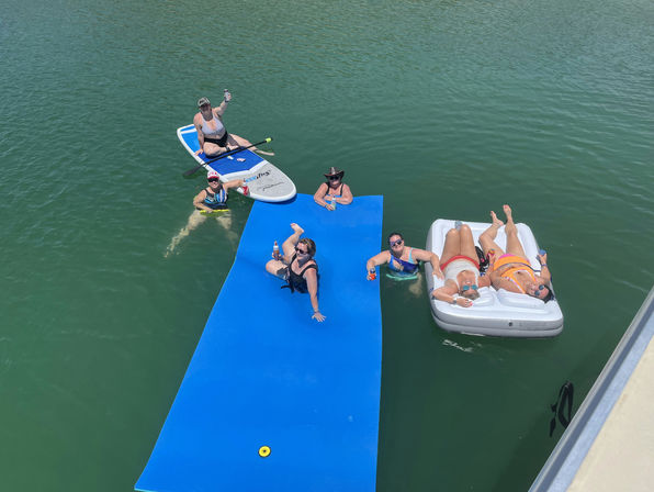 Friends floating on a bright blue water mat, a paddleboard and an inflatable lounger in a sunlit lake, sipping drinks and relaxing.