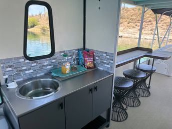 Pontoon boat kitchenette with stainless sink and mosaic tile backsplash, tray holding orange juice and a bottle plus a bag of spicy chips on the counter; three black wire‑frame bar stools at an open bar overlooking a calm lake and arid desert shoreline.