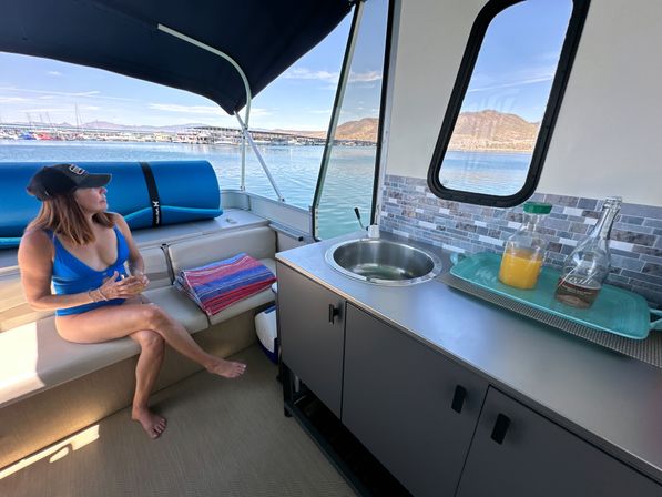 Woman in blue swimsuit and black cap relaxing on a pontoon boat bench with a drink, compact galley sink and tray of orange juice and bottles, folded towels and cooler, marina and desert mountains visible through the window on a sunny lake day.