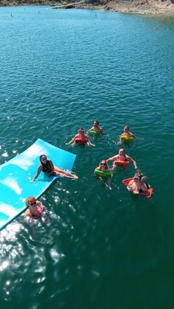 Seven people swimming and relaxing on colorful floatation devices and a large blue float mat in a turquoise lake by a rocky shoreline on a sunny summer day