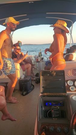 Friends enjoying a summer sunset on a pontoon boat — man in a cap holding a beer and a woman in a straw hat and bikini chatting near a table of drinks with calm lake water and distant shoreline in the background.