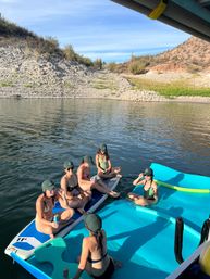 Friends in swimsuits and green caps relaxing on a paddleboard and bright blue float mat at a calm desert lake with rocky shoreline and cacti on a sunny day