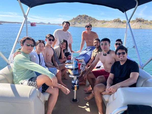 Group of people relaxing on a shaded pontoon boat on a sunny desert lake, seated around a small table with drinks and snacks and rocky shoreline in the background.