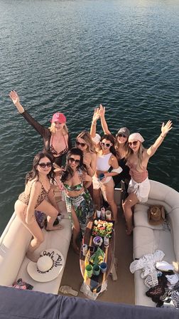 Sunny pontoon boat party on a lake with a group of women in swimsuits cheering around a snack and drink table