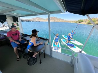 Guests on a covered pontoon boat watching three stand-up paddleboarders on clear turquoise lake with desert hills on a sunny day