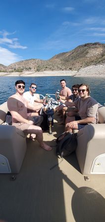 Five friends toasting canned drinks on a pontoon boat at a sunny desert lake, blue sky overhead and rocky, cactus-studded hills on the shoreline.