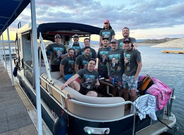 Group of people in matching graphic T-shirts smiling on a pontoon boat docked at a lakeside marina at sunset, calm water and rocky shoreline in the background.