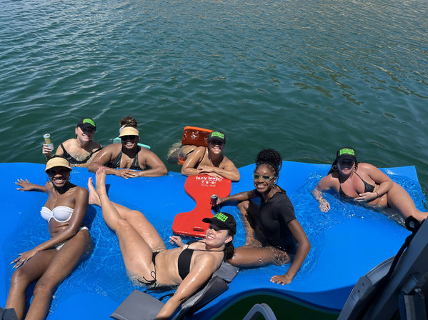 Group of women relaxing on a bright blue floating mat beside a boat on a calm lake, wearing swimsuits and caps, smiling and holding drinks on a sunny summer day.