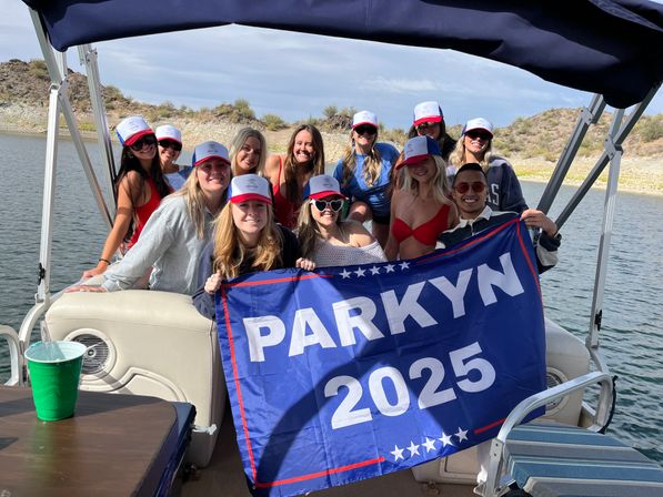 Friends on a pontoon boat at a sunny lake with rocky shoreline, wearing matching trucker hats and smiling while holding a blue "PARKYN 2025" flag for a fun boat party.