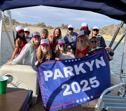 Friends on a pontoon boat at a sunny lake with rocky shoreline, wearing matching trucker hats and smiling while holding a blue "PARKYN 2025" flag for a fun boat party.