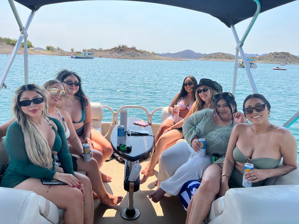 Group of eight women in swimsuits smiling and holding canned drinks on a shaded pontoon boat cruising a blue lake with rocky shoreline and low hills on a sunny summer day.