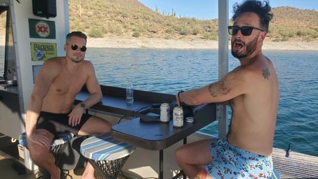 Two shirtless friends in sunglasses chilling on a pontoon boat at a sunny desert lake with cacti-studded hills, drinks on the table and a water bottle nearby.