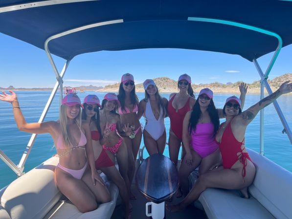 Eight women in colorful swimsuits and matching pink caps posing on a covered pontoon boat over a calm blue desert lake with rocky, cactus-dotted hills and a clear sky.