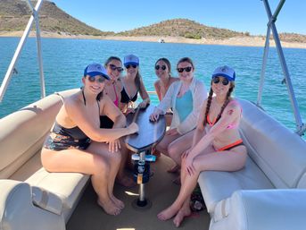 Six women in bikinis and blue caps smiling around a table on a pontoon boat on a sunny lake with rocky desert hills in the background