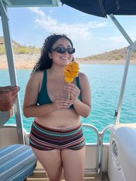 Smiling woman in sunglasses and a bikini top on a pontoon boat holding a spiral-cut mango on a stick, with turquoise lake and rocky, cactus-studded shore behind her.