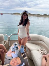 Smiling woman in a white swimsuit wearing a captain’s hat aboard a pontoon boat on a calm lake, extending an arm with a small novelty face sticker; a wooden table holds oversized face cutouts on sticks and light-blue tumblers, with shoreline and cloudy sky in the background.