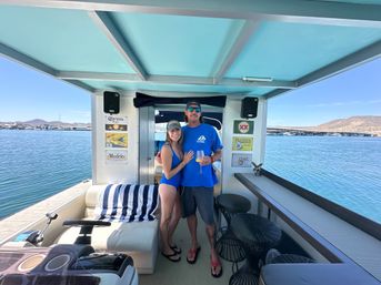 Cheerful couple in swimwear on a covered houseboat deck holding drinks by a striped lounge, overlooking calm blue water, marina docks and rolling hills under a clear sky.