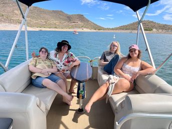 Four people relaxing on a pontoon boat on a sunny desert lake, blue water with cactus‑dotted hills and a clear sky in the background.