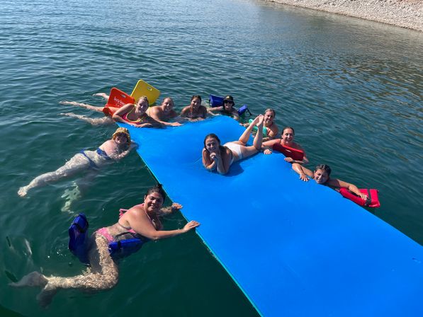 Group of swimmers lounging on and around a bright blue floating mat in clear lake water near a pebbled shoreline on a sunny summer day