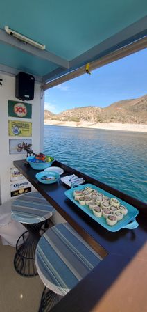 Lakeside boat bar counter with turquoise platter of pinwheel wraps, bowls of snacks and striped stools overlooking a sunny desert lake and arid hills.