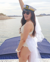 Bride-to-be in a captain's hat and veil on a boat at a lake, wearing a white swimsuit and sunglasses, smiling with one arm raised against a rocky hilly shoreline