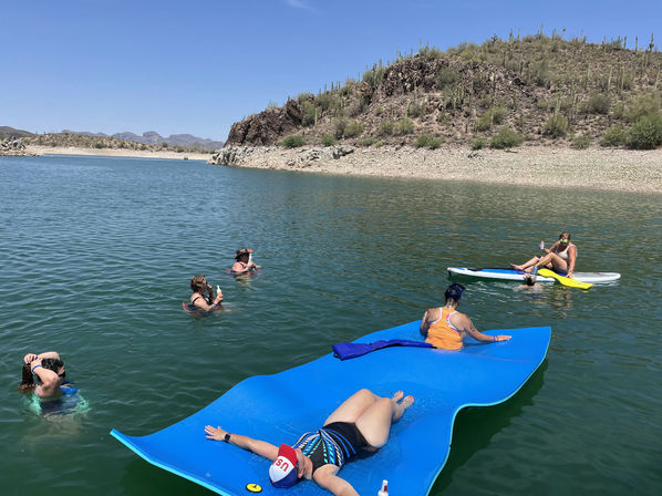 Friends relaxing on a bright blue floating mat and paddleboards on a calm desert lake with rocky shoreline and saguaro-studded hills under a clear blue sky — summer water recreation.