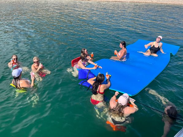 Group of friends relaxing on a bright blue floating mat and in clear lake water near a pebbled shore, holding drinks on a sunny summer day